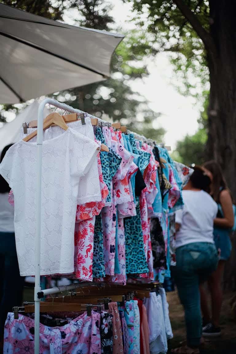 Colorful shirts hanging on a rack at an outdoor market with people browsing.