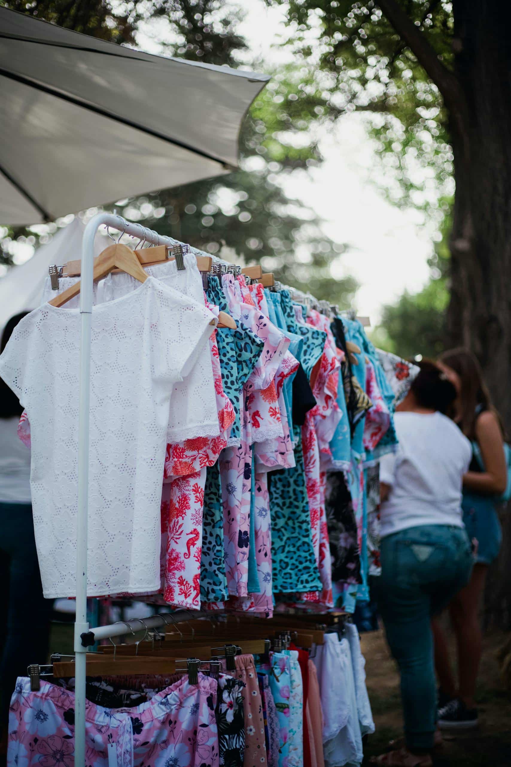 Colorful shirts hanging on a rack at an outdoor market with people browsing.