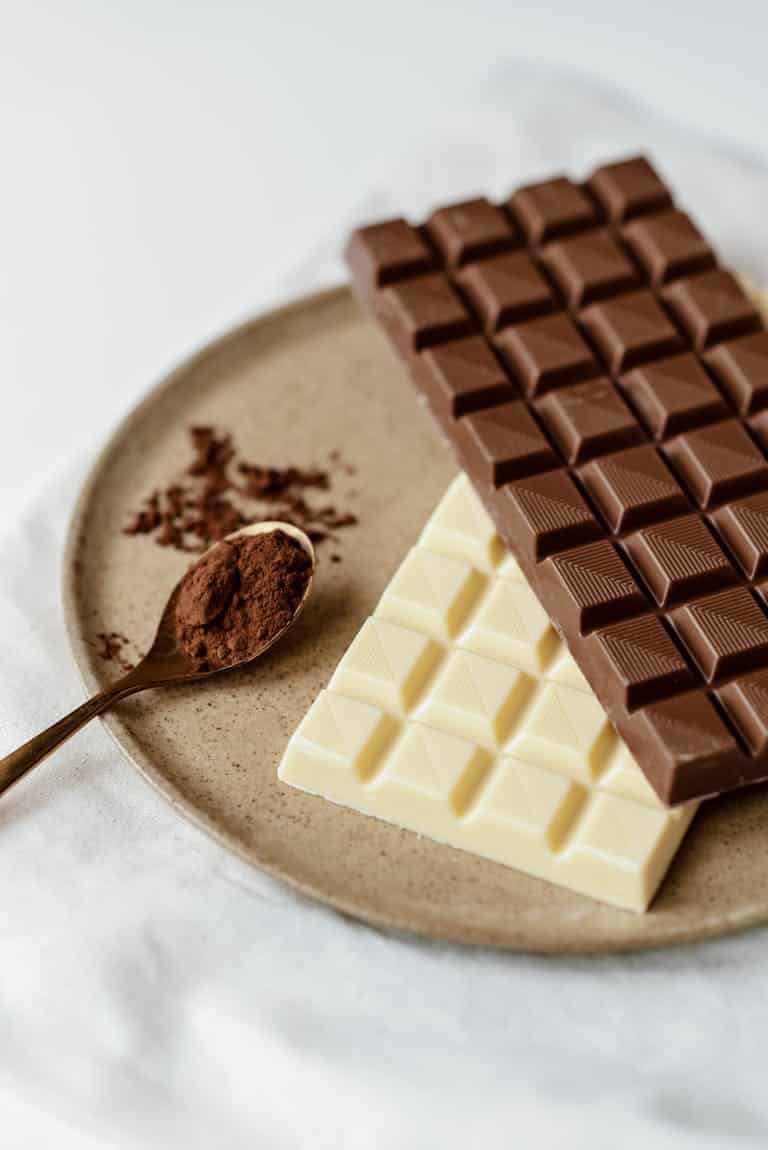 High angle of delicious milk and white bars of chocolate on ceramic plate near spoon with cacao powder on white textile