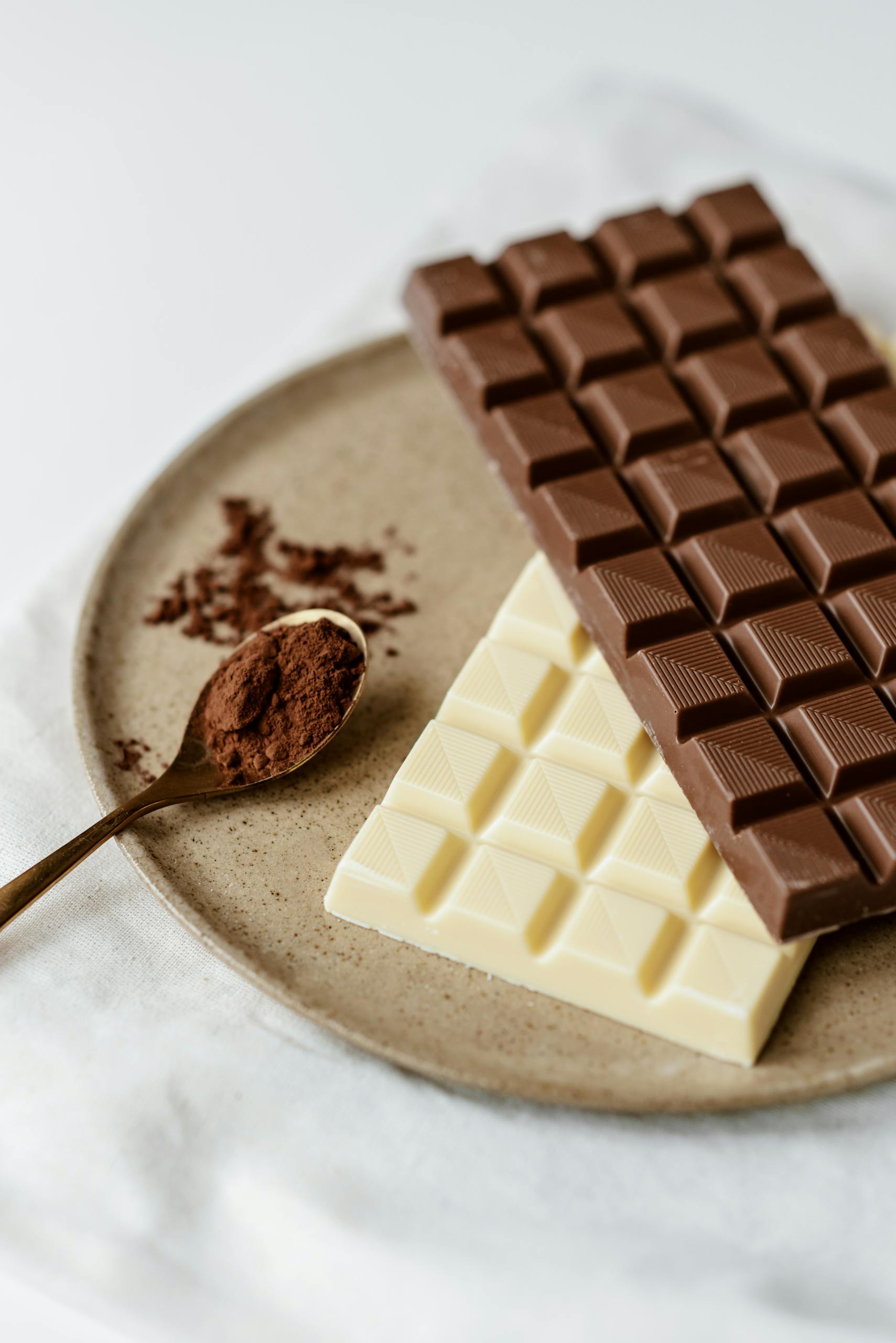 High angle of delicious milk and white bars of chocolate on ceramic plate near spoon with cacao powder on white textile