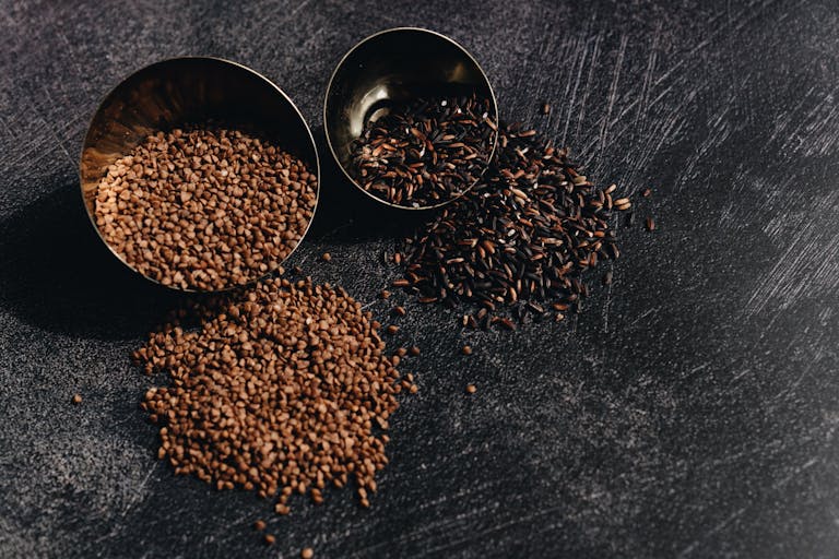 Top view of raw buckwheat and wild rice in bowls on a dark textured surface.