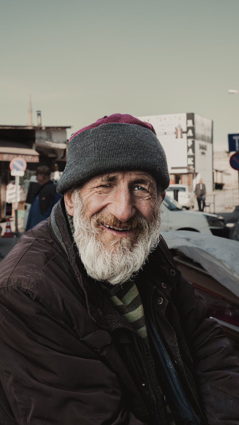 A smiling elderly man with a grey beard and hat in an urban outdoor setting.