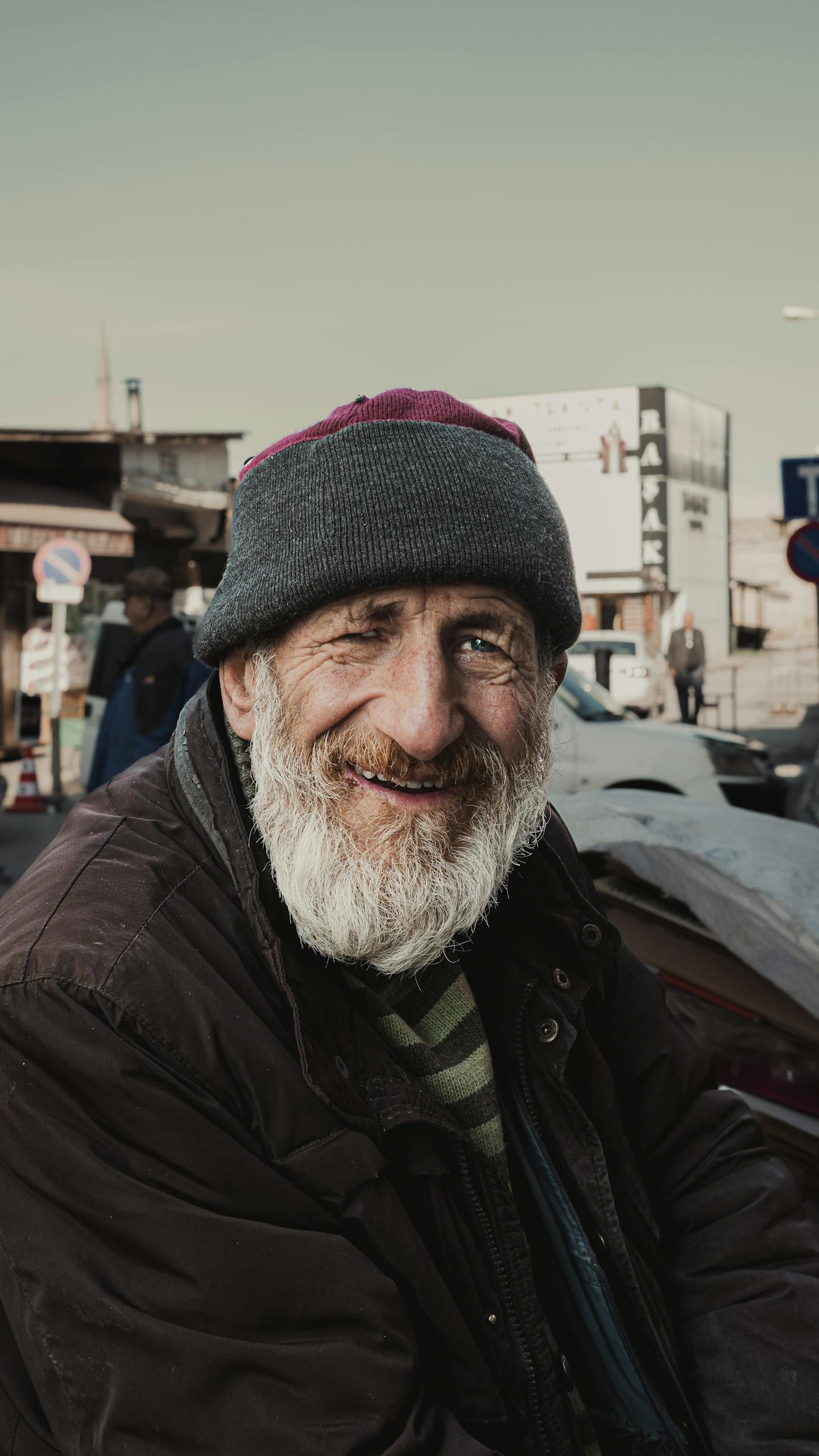 A smiling elderly man with a grey beard and hat in an urban outdoor setting.
